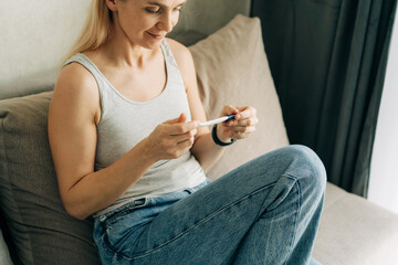 A woman is holding a pregnancy test in her hands and waiting for the result sitting on the couch.