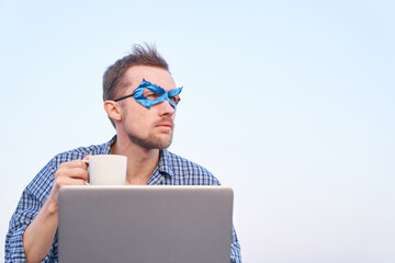 IT support superhero guy using laptop holding cup of hot tea or coffee. Cheerful man in nightwear shirt and blue face mask using laptop outdoors. High quality image