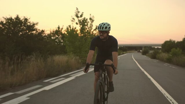 Happy bearded man in protective helmet, mirrored glasses and sportswear riding a bicycle on an asphalt road outside the city in the evening.
