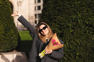 Naklejka premium Happy attractive woman hold bouquet pink flowers walk outdoors, meeting her friends or love, showing high five hey hello hi sign. She is joyful to meet her acquainted by chance. Girl wave her hand.