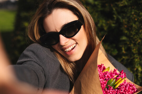 Close up portrait of attractive blonde woman take selfie on her mobile phone. Woman wear sunglasses, grey jacket and hold pink flowers bouquet. Outdoor selfie in fashion outfit while walking in park.