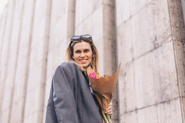 Fototapeta na wymiar Positive girl hold pink flowers in the city center. Amazing blonde haired lady with natural make up look happy, walking outdoors in grey jacket and hold pink bouquet in craft paper, girl look happy.