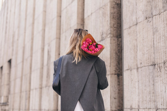 Back view image of a beautiful amazing young blonde woman posing while walking outdoors and holding bouquet of pink flowers on shoulders. Girl walking on the street and wear grey jacket.