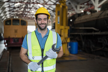 Male engineer worker working in industry factory, standing with smile and holding industrial spanner, wearing safety uniform and helmet. Male technician worker at work in locomotive garage