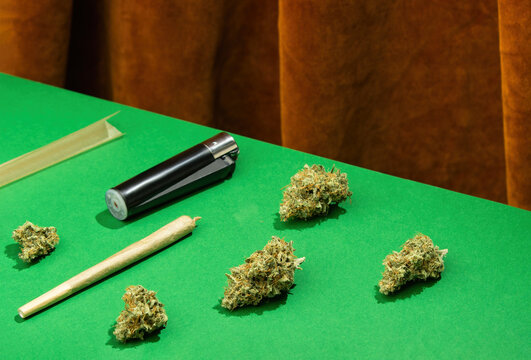 King size joint, paper and lighter lie among dry marijuana buds on a green table against a background of brown velvet curtains