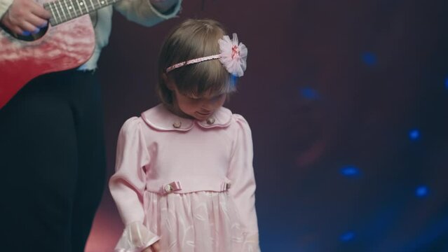 Little girl on stage in pink vintage dress, she dances, her father plays acoustic guitar. Color music is shining. Performance on stage. Early development of children
