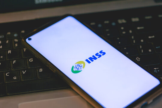 A logo do INSS na tela de um telefone celular que est&aacute; sobre um teclado de um laptop. Instituto Nacional do Seguro Social.