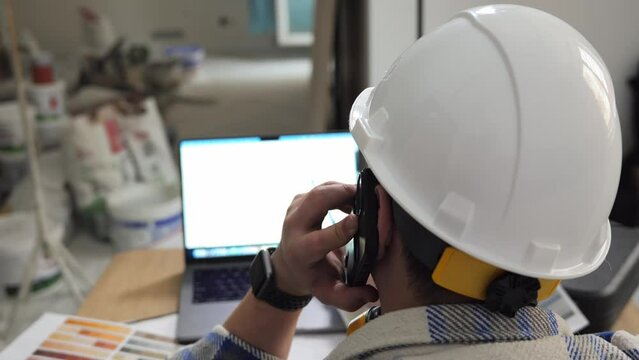 A view from the back of a construction engineer on a construction site indoors, who is talking on the phone and looking at a drawing of the room on a laptop. An unrecognizable person. High quality 4k