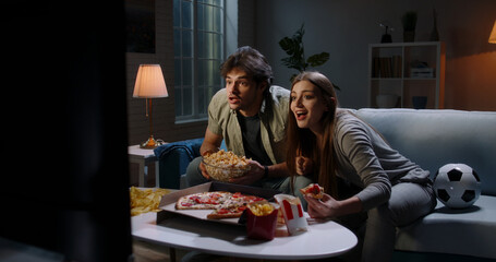 Funny young emotional caucasian sport fan couple watching a game together, eating popcorn and pizza and reacting to their team playing 