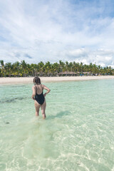 An anonymous female tourist in a black one piece swimsuit enjoys the clear waters of a tropical beach in the summer.
