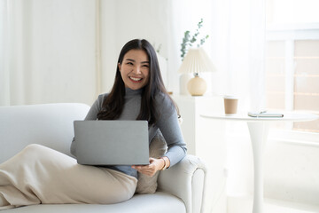 Young beautiful Asian woman using laptop computer while sitting on the sofa at her living room.
