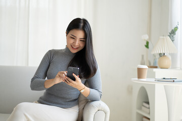 Young attractive Asian female using smartphone while sitting on the sofa in living room.