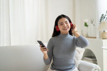 Happy young Asian woman wear earphones relax on couch listen to music on smartphone.