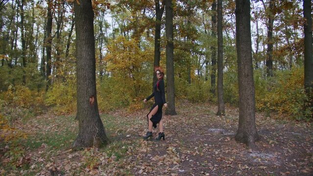 A young woman with Santa Muerte make-up dressed in a black dress of death walks against the backdrop of autumn leaves in the forest during sunset. Day of the Dead or Halloween concept.