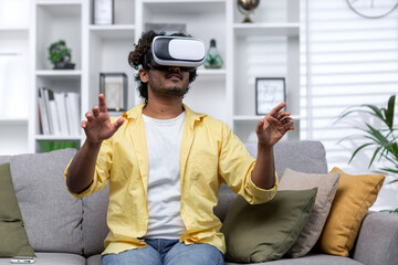 Latin American man sitting on couch at home using virtual reality glasses, man viewing educational material remotely.