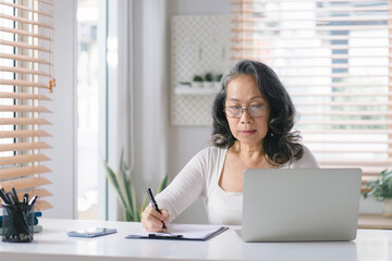 Senior stylish entrepreneur wearing eyeglasses, working on a computer, with a notebook and pen at her home office.