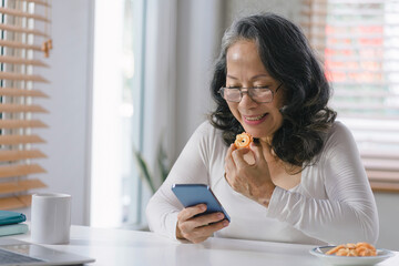 Obraz premium Capturing the Joys of Aging: A Happy Senior Woman Embracing Technology as She Navigates Her Smartphone with Ease from the Comfort of Her Home.