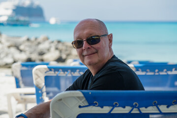 A bald, senior gay man wearing sun glasses and a black t-shirt, sits sideways on a beach chair and...