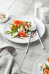 Healthy vegetable salad with fresh arugula, tomato, feta cheese on white plate with cutlery, gray tile background. Diet menu with text space