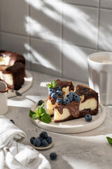 Two pieces of homemade chocolate cheesecake with blueberries, chocolate sauce and mint on a plate with coffee on white kitchen table for morning breakfast in hursh light. Text space, copy space