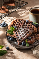 Heart form chocolate waffle cookies in ceramic plate decorated with berries, powdered sugar and mint with tea on beige background. Sunny morning breakfast with hursh shadows
