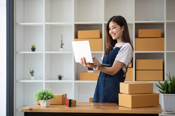 Obraz na płótnie Canvas A portrait of a young Asian woman, e-commerce employee sitting in the office full of packages in the background write note of orders and a calculator, for SME business ecommerce and delivery business.