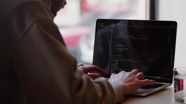 A freelance programmer works at a computer while sitting in a cafe. A male developer writes code for a website. View from behind.