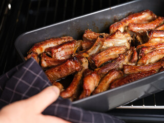 A woman hand taking a black baking tray with baked pork ribs out of an oven.