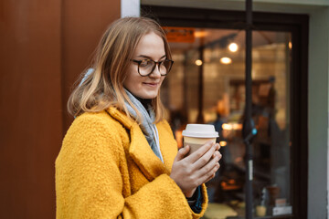 Charming happy young woman dressed in a bright yellow coat holds coffee to go. Portrait of smiling girl in glasses spend free time traveling in european old town enjoying cup of tea, coffee. 