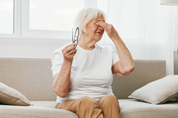Elderly woman severe eye pain sitting on the couch, health problems in old age, poor quality of...