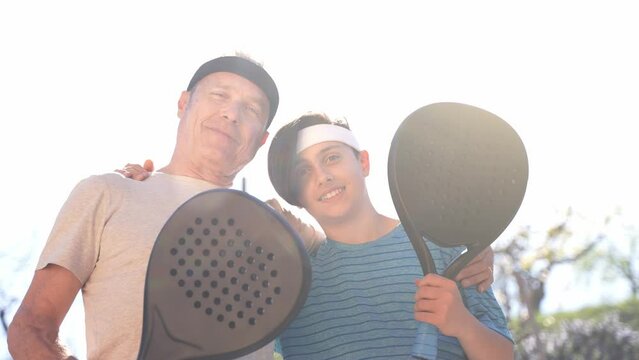 Backlit shot of a happy smiling grandfather or father and grandson or son holding padel rackets and embracing each others outdoors.