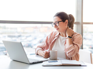 Woman winces from pain in her neck. Female office employee tries to stretch stiff neck muscles while working with laptop. Modern office at co-working center. Workplace for freelancers or students.