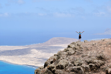 Obraz premium Woman with arms raised on top of a hill. With La Graciosa Island in the background