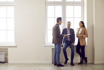 Full-size portrait of attractive smiling colleagues standing near bright window in spacious room....