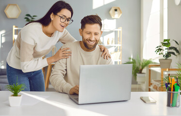 Happy young family couple using a laptop. Loving, supportive woman standing by her husband who is working on his notebook at his desk, holding him by the shoulders, looking at the computer and smiling