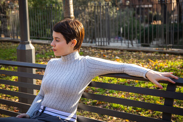 The young woman is sitting on a park bench in Rome. Beautiful woman with short hair is relaxing in the park.