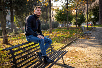 The young man is sitting on the bench in the park in Rome. Beautiful man in a trendy suit is waiting for his girlfriend. - 585168885