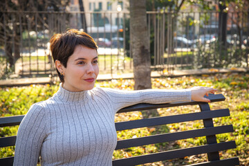 The young woman is sitting on a park bench in Rome. Beautiful woman with short hair is relaxing in the park.