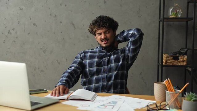 Young sad tired employee IT Indian business man wear casual clothes blue shirt sit work at office desk with laptop pc computer hold neck make head tilts suffer from pain Professional staff job concept