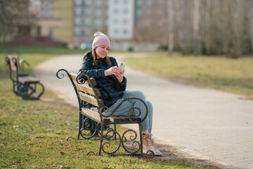 Portrait of woman with smartphone in hands sitting on wooden bench at city street in spring day