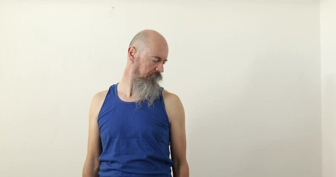 50 years old man with long beard doing neck and upper spine exercise at home, banding head sides towards shoulders over white wall
