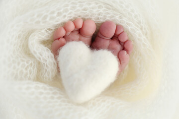 Knitted white heart in the legs of a baby. Macro photography. The tiny foot of a newborn baby. Soft feet of a new born in a white wool blanket. Close up of toes, heels and feet of a newborn.