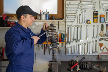 Image of a mechanic in overalls in his workshop while he disassembles a car shock absorber with a wrench and extractors.
