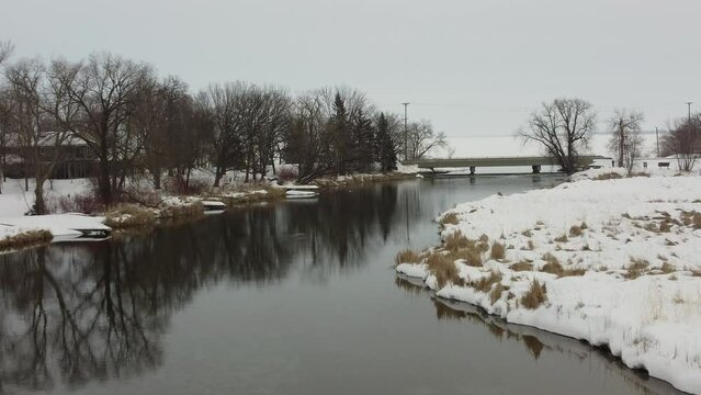 Aerial camera moves upstream along a placid river toward a dam in winter