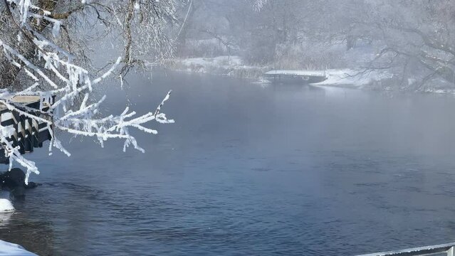 Fog rises from a clear river flowing near hoar frost covered trees