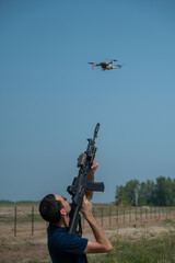 A man aims to shoot a rifle at a flying drone against a blue sky. 