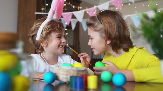 Easter Family traditions. Two caucasian happy siblings kids with bunny ears paint and decorate eggs with paints for holidays while sitting together at home table. Kids having fun together. 