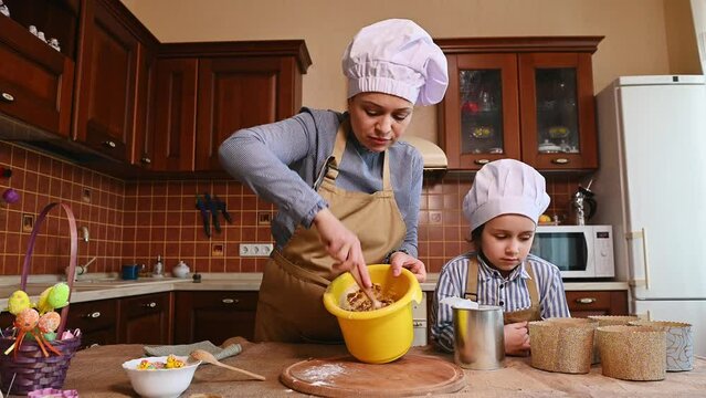 Middle-aged multi-ethnic woman, loving mother and her lovely daughter in chef's hats and aprons, standing by a kitchen island, kneading dough, cooking together festive bakeries for Easter holidays