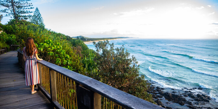 A girl in dress walks along the boardwalk and enjoys the view of the stunning Coolum Beach and surfers catching big waves, Pacific Ocean. Stunning panorama of Sunshine Coast, Queensland, Australia