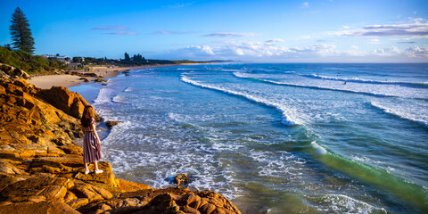 A beautiful girl in a dress stands on a rocky cliff looking at the famous Coolum beach and the Pacific Ocean and surfers catching big waves. Panorama from Sunshine Coast, Queensland, Australia - obrazy, fototapety, plakaty